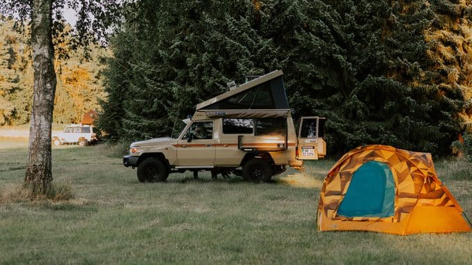 Beiger Geländewagen mit Aufstelldach und ein orangefarbenes Zelt auf einer Wiese am Waldrand